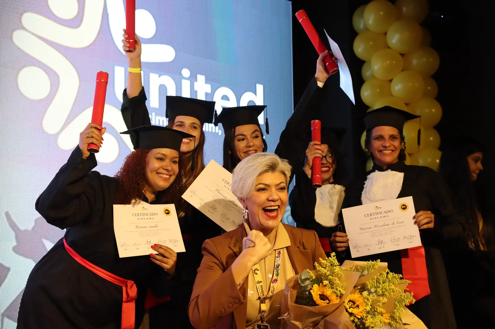 Formandas da Universidade de Vendas erguendo os certificados no palco, com a equipe da United