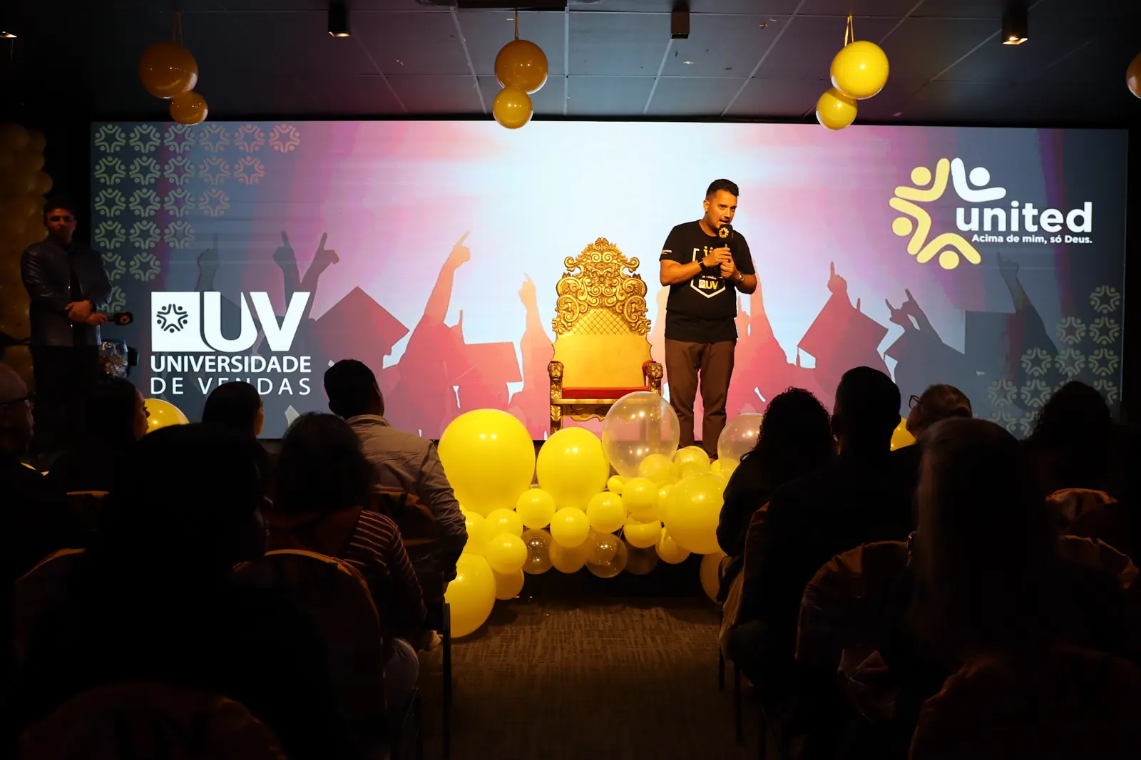Palco do Centro de Convenções com os logos da Universidade de Vendas e da United, balões amarelos e a plateia em silhueta